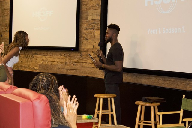 Tim Salau and Krishna Lyles stand in front of an audience with a stool in front of them.