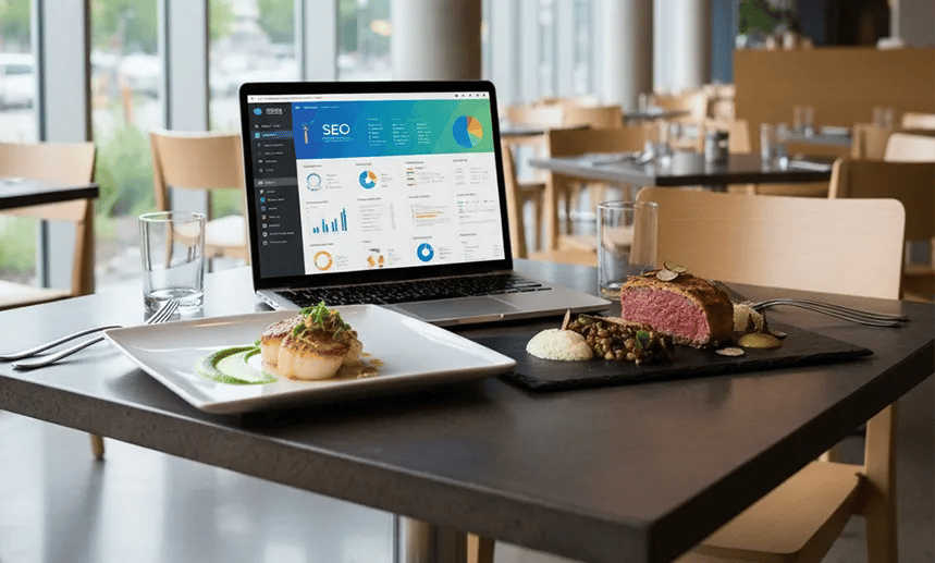 Mesa de restaurante com pratos gourmet e notebook exibindo gráficos de SEO, representando o marketing digital gastronômico.
