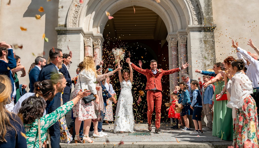 A happy bride and groom exiting a stone church while wedding guests throw colorful confetti.