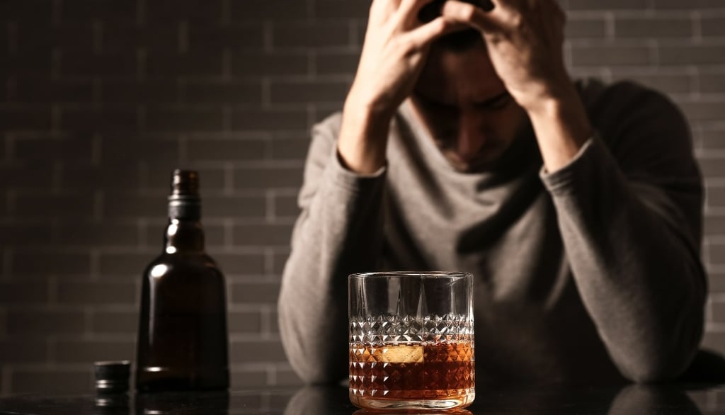 A distressed man with his head in his hands sits at a table in front of a bottle and a glass of liquor