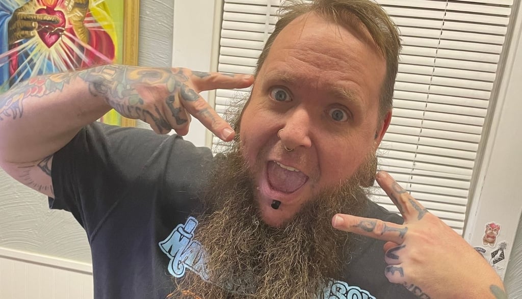 Fecke poses in his piercing room, making peace signs, an excited face, long beard, septum & lip jewelry, & tattooed hands.
