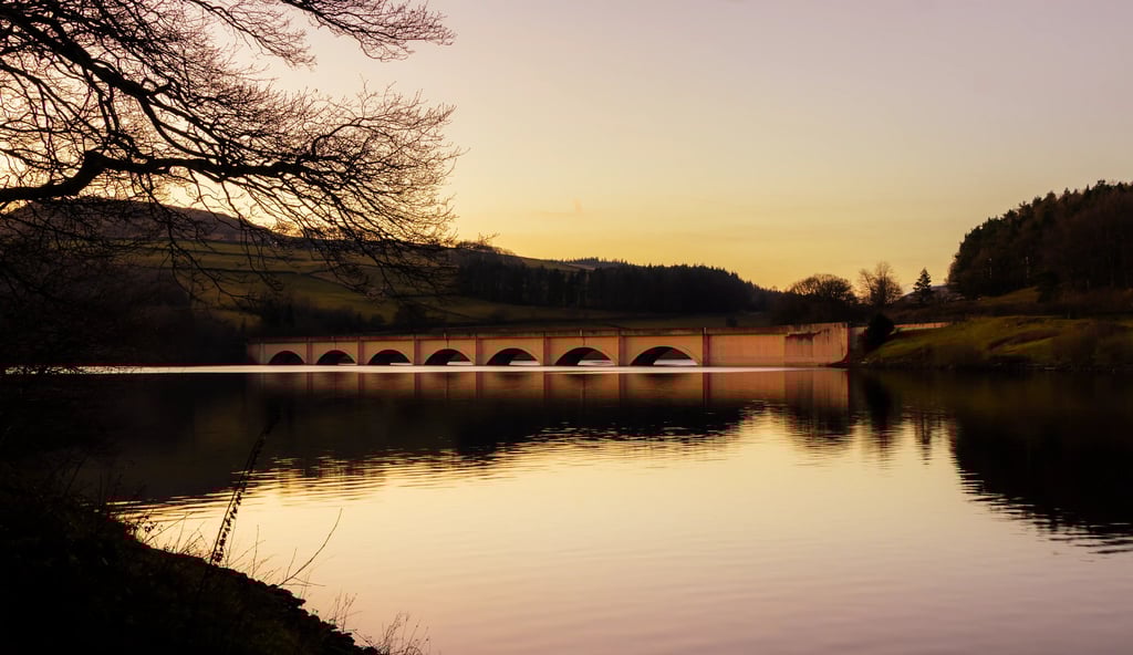 Arched stone bridge reflecting in a calm lake at sunset in a rural landscape.