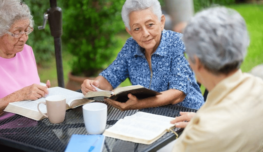 Three elderly women engaged in a book club meeting outdoors, exemplifying senior participation in li