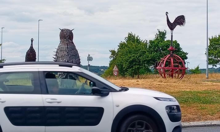 Metal animal sculptures including an owl and rooster on a grassy roadside roundabout with a passing white car.