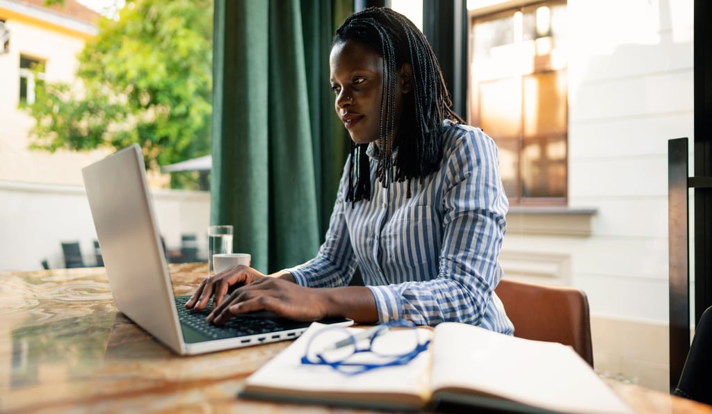 A focused black woman with braids working on a laptop at a cafe table with a notebook.