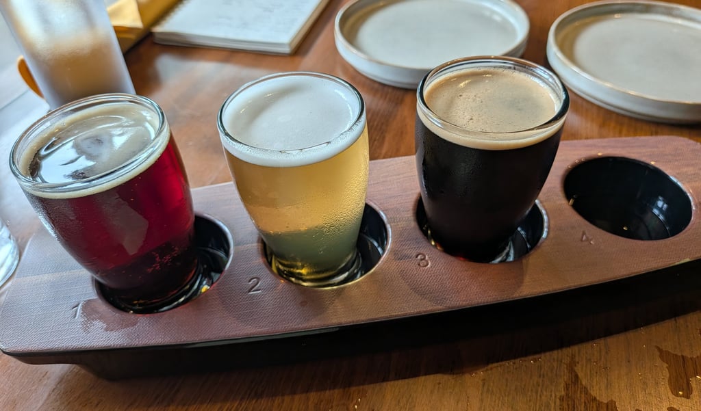 A craft beer flight with red, blonde, and dark stout samples on a wooden tasting paddle.