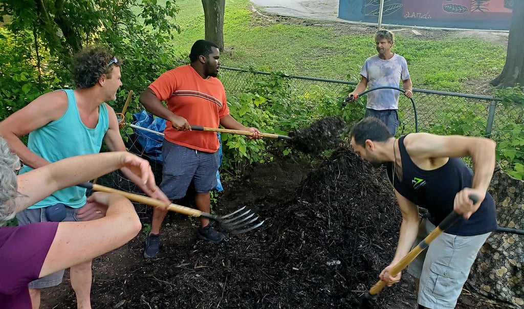 Diverse group of community volunteers using shovels to turn a large compost pile in a public park garden.