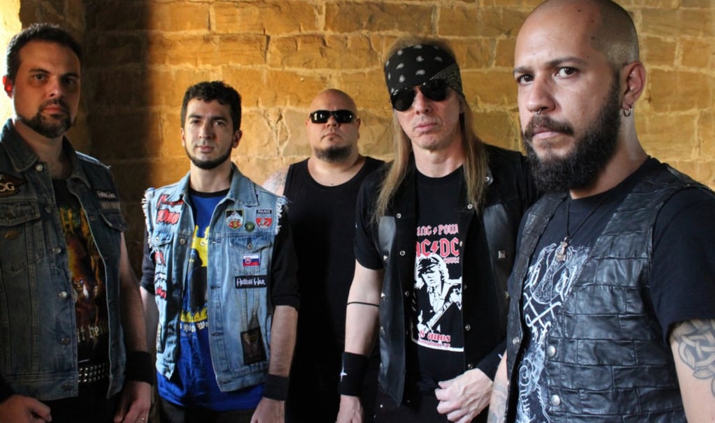 Heavy metal band members wearing denim vests and band t-shirts posing against a stone wall.