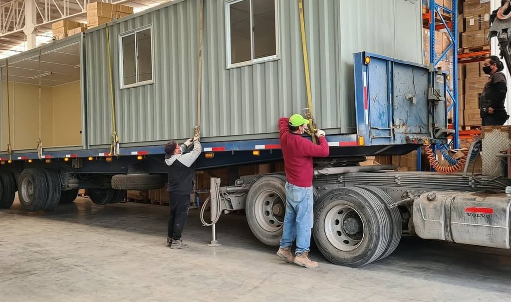 Workers securing a gray prefabricated container office onto a flatbed semi-truck for transport inside a warehouse.