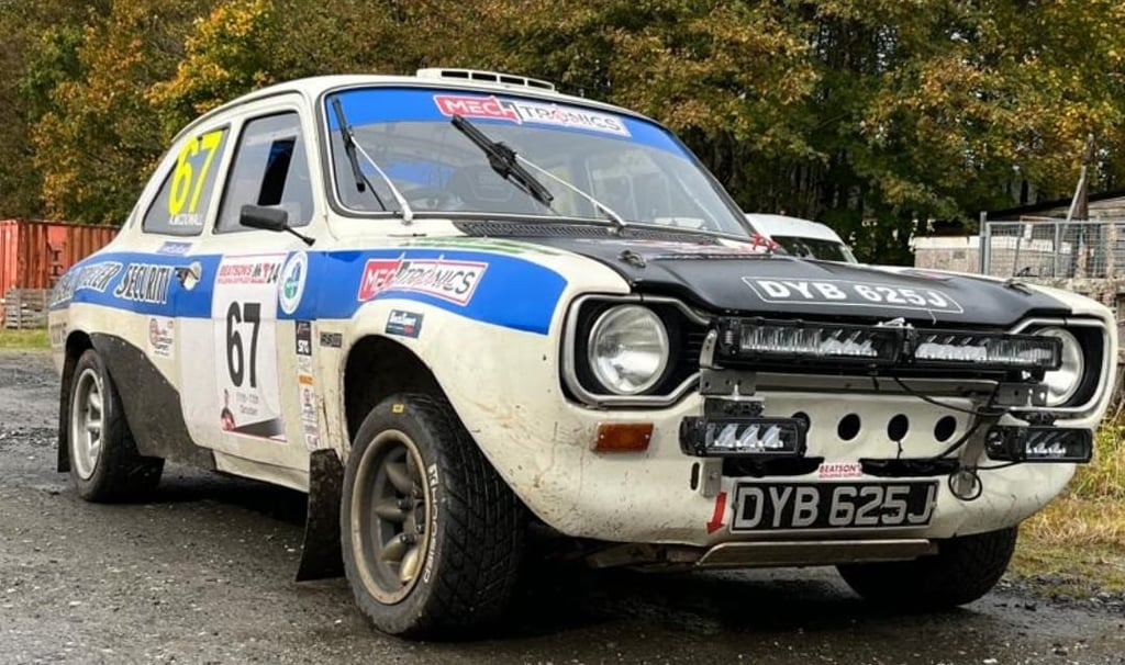 White Ford Escort MK1 rally car with sponsor decals and auxiliary light pods parked on a gravel track.