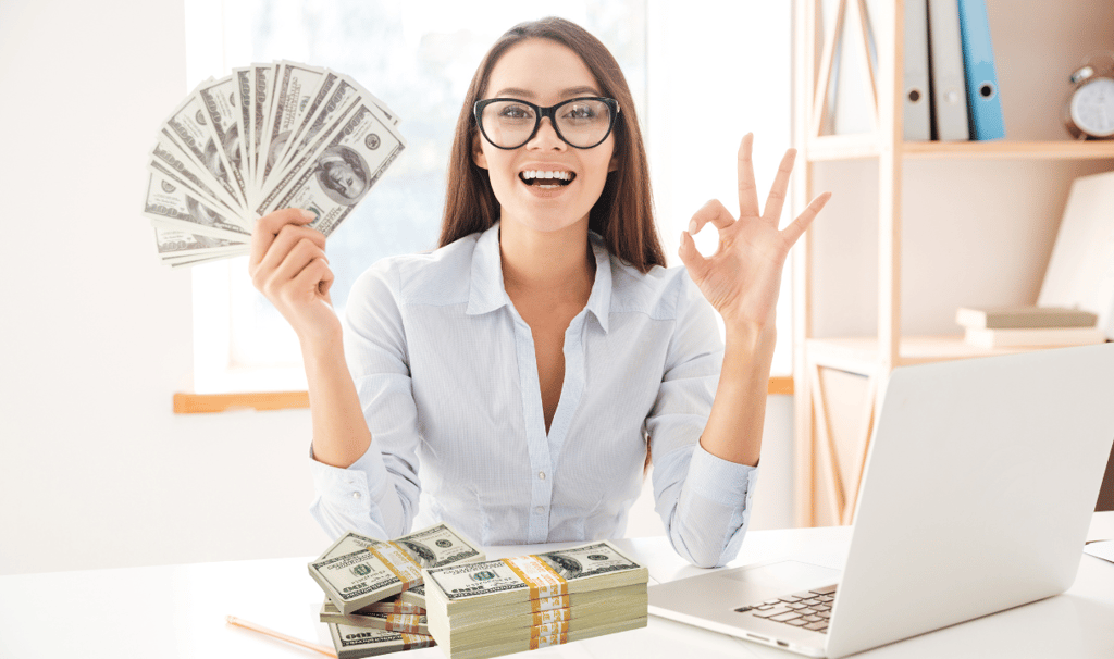 a woman sitting at a desk with a laptop and holding up a stack of money