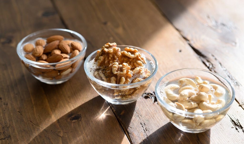 Bodegón de diferentes cuencos de cristal con almendras, nueces y anacardos sumergidos en agua sobre una mesa de madera clara