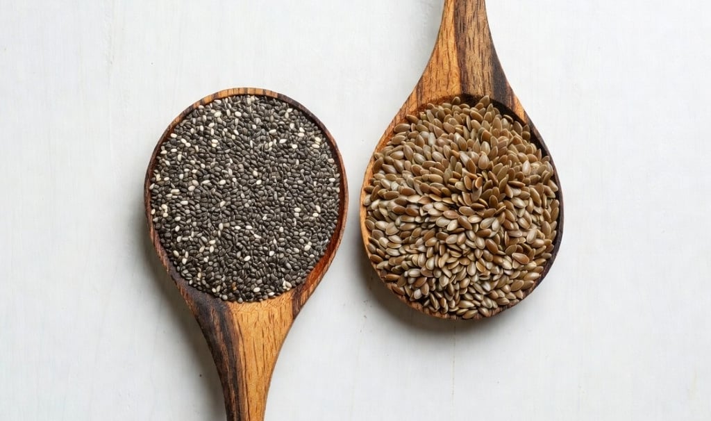 Dos cucharas de madera cruzadas sobre una mesa, una llena de semillas de chía negras y otra de semillas de lino dorado