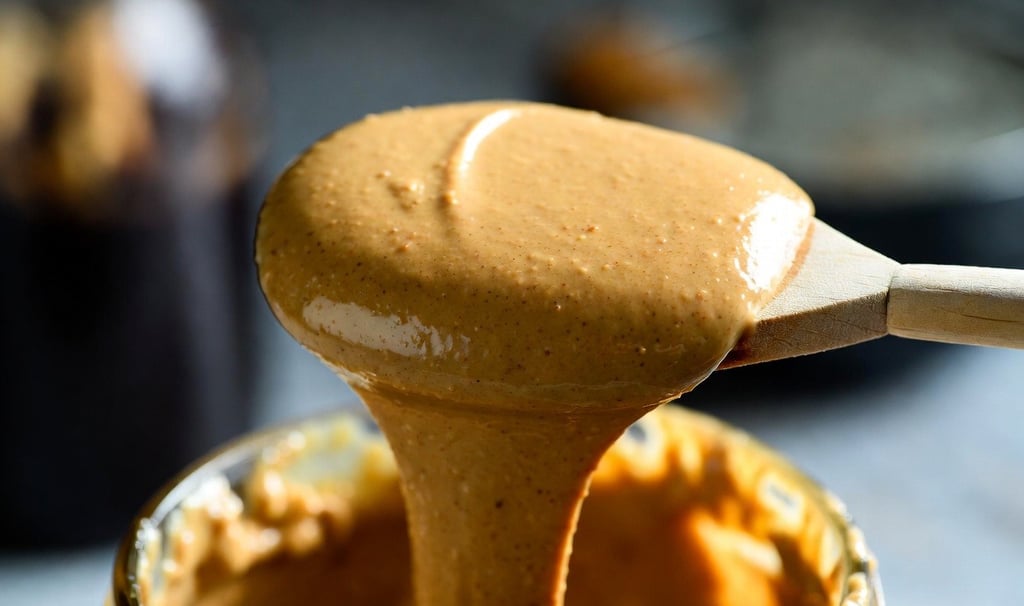 Primer plano de una cuchara de madera goteando mantequilla de almendras cremosa sobre un tarro de cristal - Raw Rico