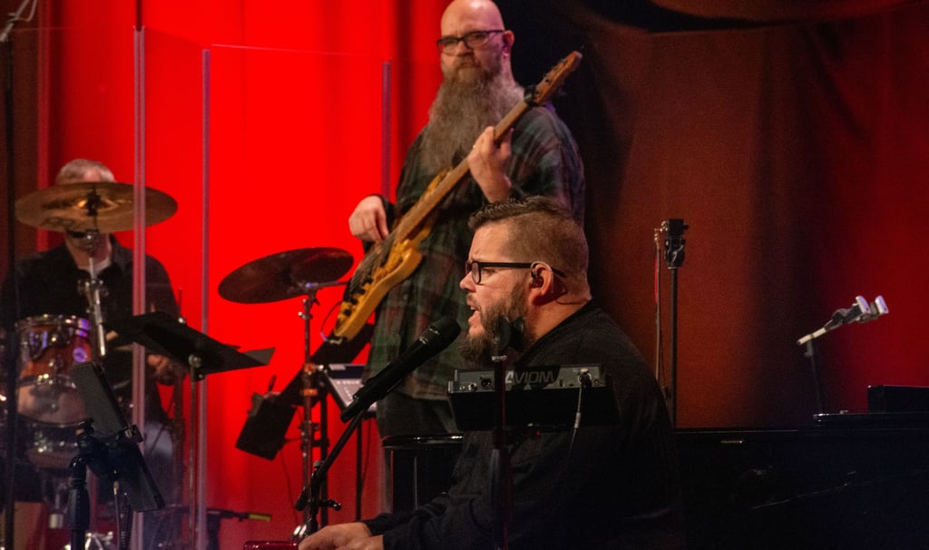 A worship team playing music on a church stage, with drums, bass guitar, piano, and vocalists