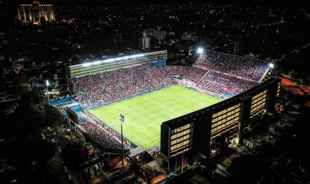 Vista panorâmica interna do Estádio Gran Parque Central com arquibancadas em azul, branco e vermelho sob luz natural.