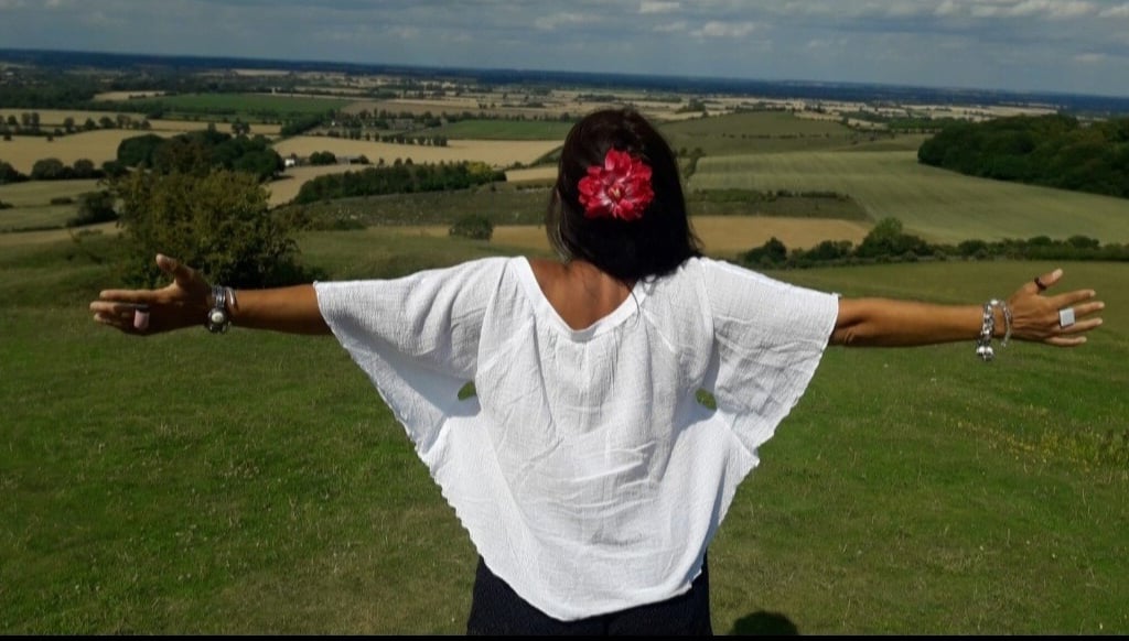 An image of the back of Sheila Cristina on the top of a hill. In front of her is a beautiful view of many fields in England.