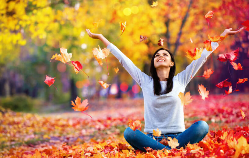 a woman joyfully sitting on a leafy field with her arms up.