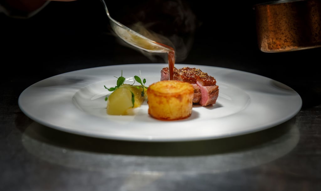 Chef pouring red wine jus over gourmet pan-seared duck breast with potato fondant and garnish.