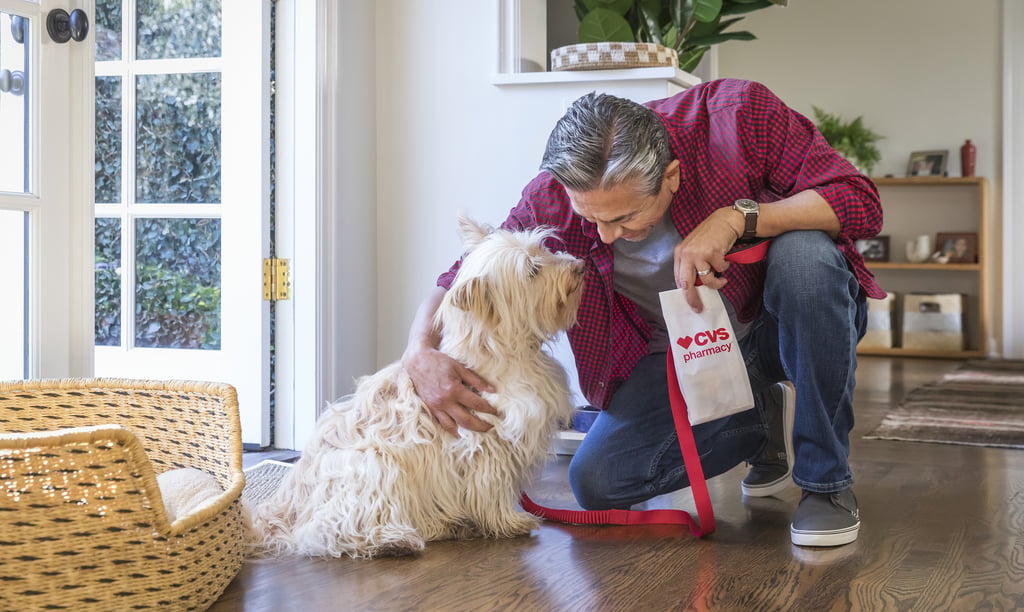 A man at home petting his fluffy dog while holding a CVS Pharmacy prescription bag for pet medication.