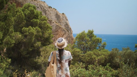 A woman in a sun hat looking at the blue Mediterranean Sea from a rocky cliff with green pine trees.
