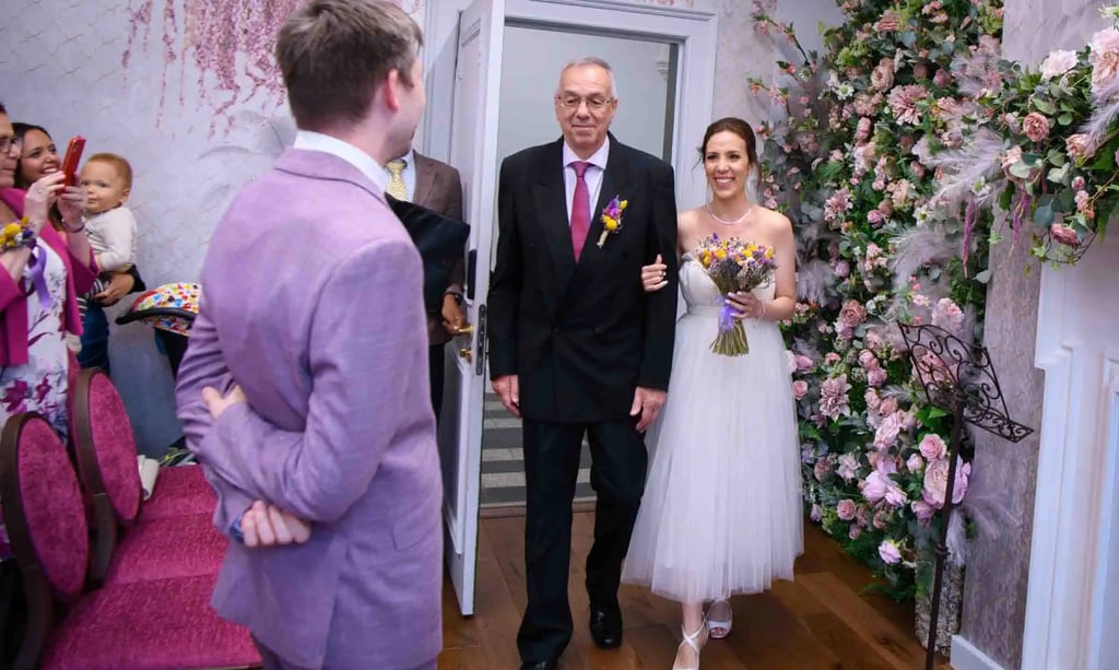 A happy bride in a white dress walks down the aisle with her father past a pink floral wall.