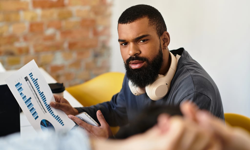 A male data analyst with a beard and headphones reviewing financial charts and business reports in a modern office.