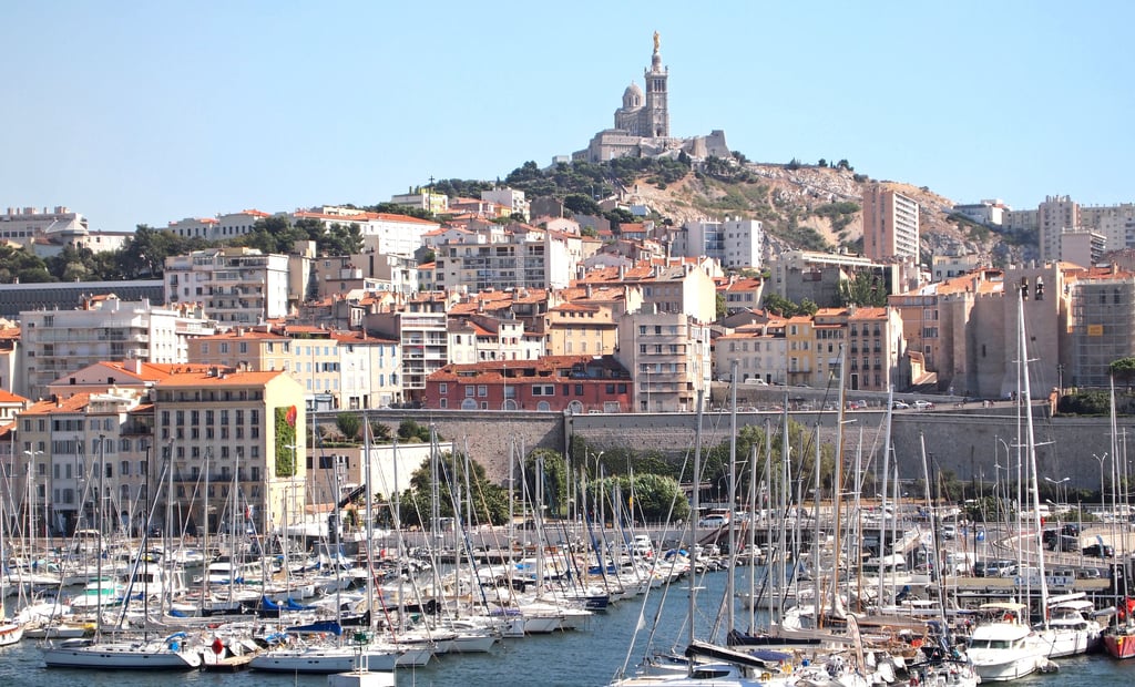 Blick auf Marseille vom Meer aus mit Fischerbooten im Vordergrund, die Altstadt und der Hafen sichtbar, im Hintergrund die Ba