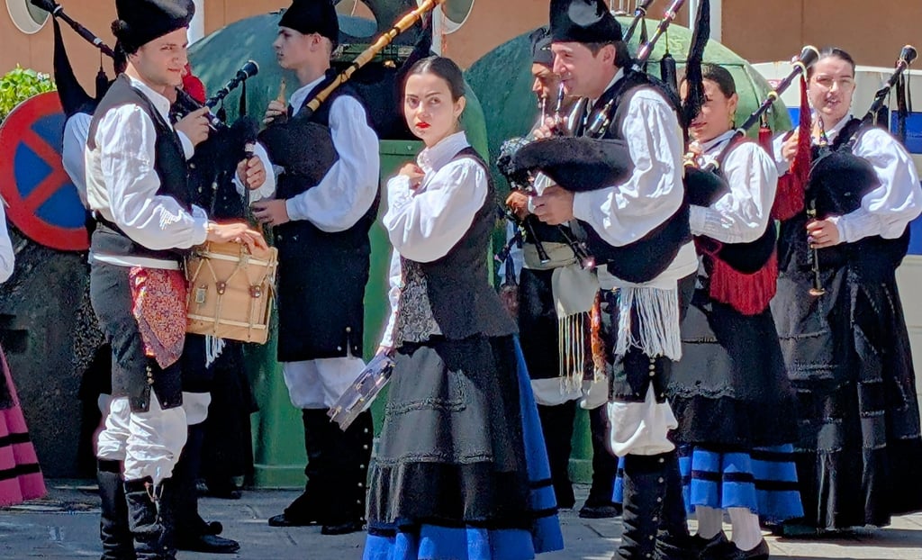 A group of musicians in traditional black and white folk attire play bagpipes and drums in an outdoor plaza in Cuntis.