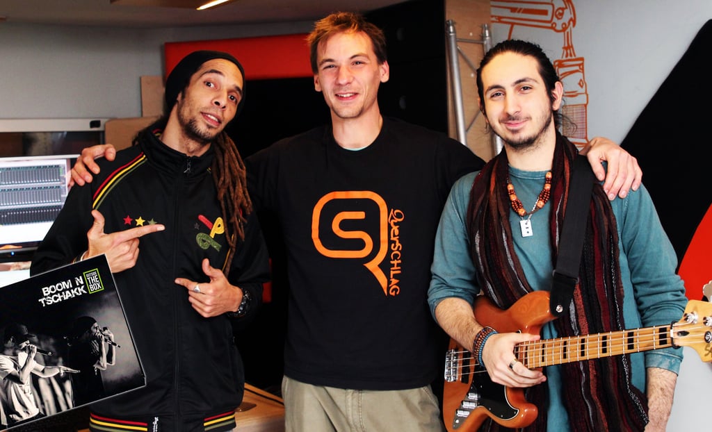 three men standing next to each other producing songs , one is holding a guitar