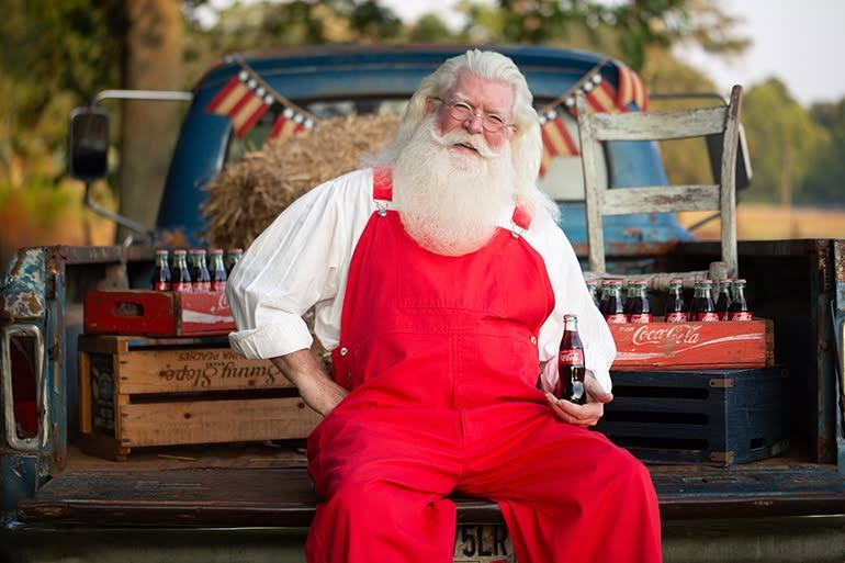 A vintage Santa Claus in red overalls holding a Coca-Cola bottle while sitting on a classic blue truck.
