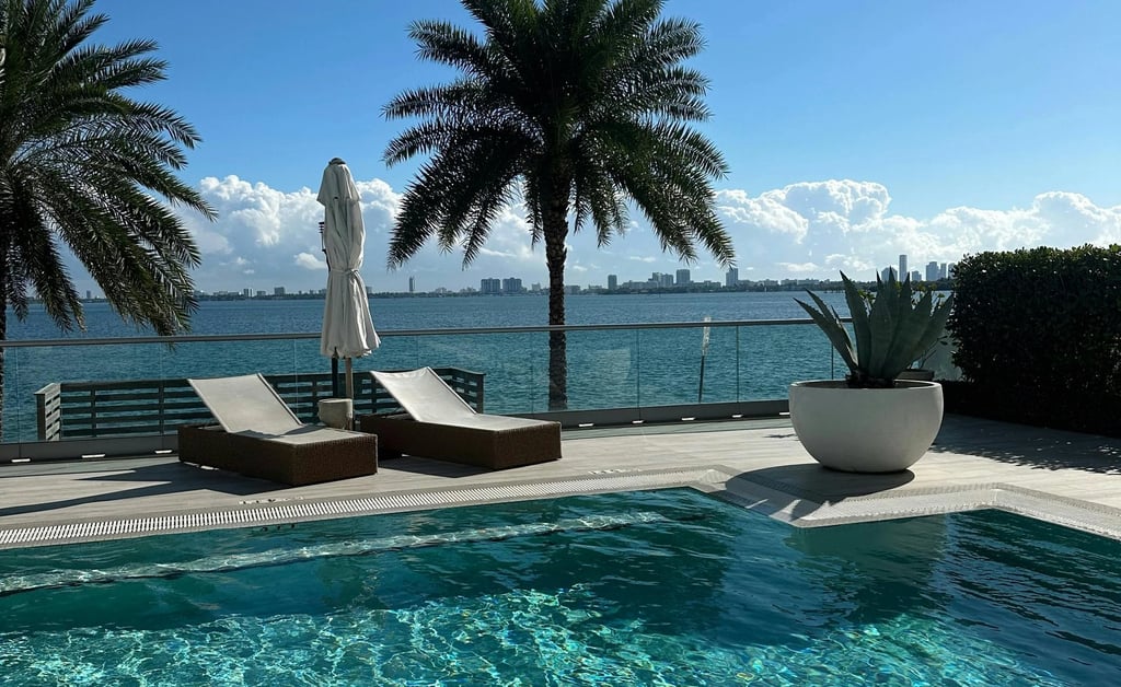 Luxury infinity pool with lounge chairs and palm trees overlooking a tropical bay and city skyline.
