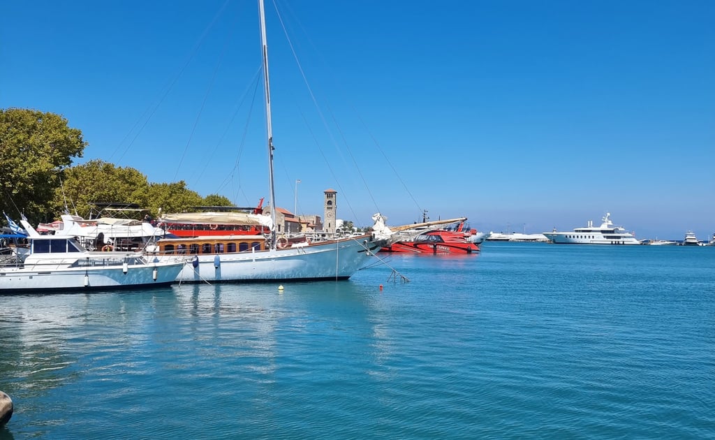 Luxury yachts and sailboats anchored in the turquoise waters of Rhodes harbor under a clear blue sky.