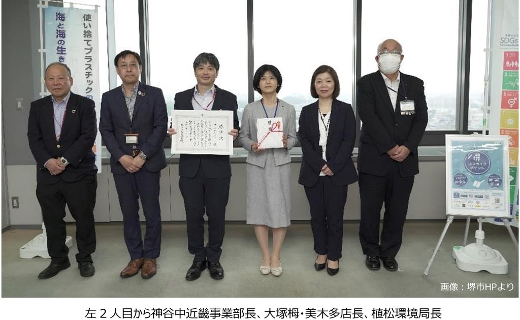 堺市役所で感謝状を受け取るダイエー関係者と堺市職員の集合写真。中央で感謝状と目録を持ち、プラスチック削減やSDGsの掲示が背景に写る。