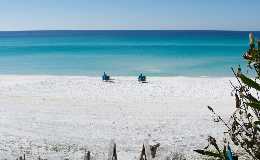 A wooden boardwalk leads to a white sand beach with turquoise ocean water and beach chairs.