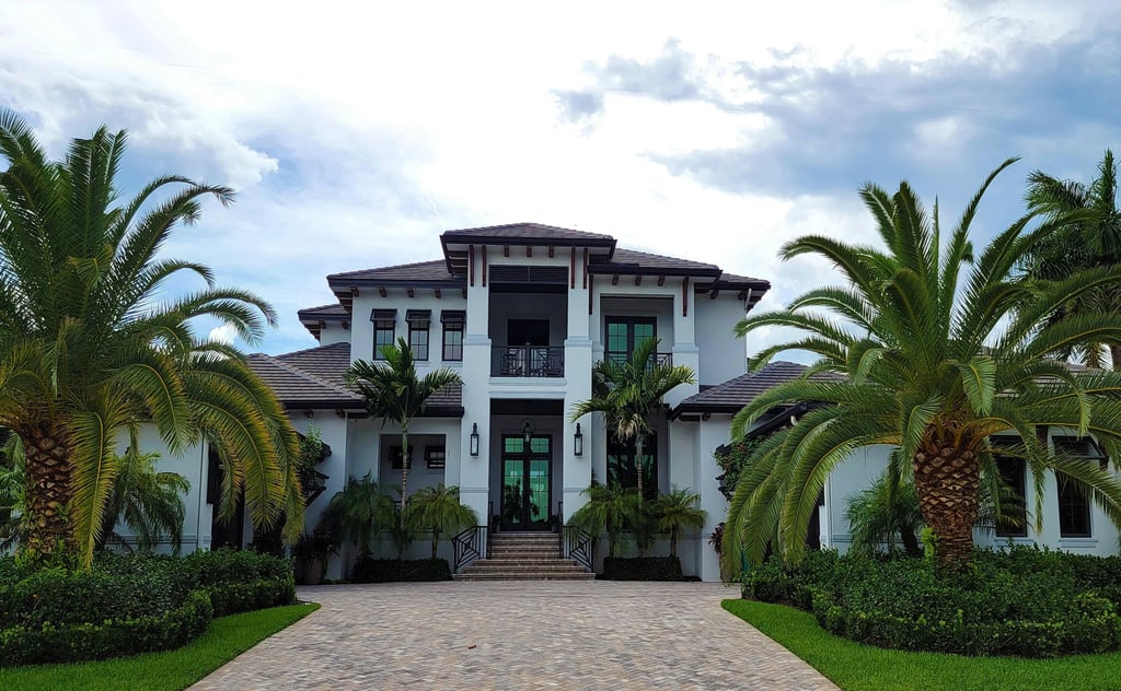Luxury white two-story coastal mansion with palm trees and a paved driveway under a cloudy sky.