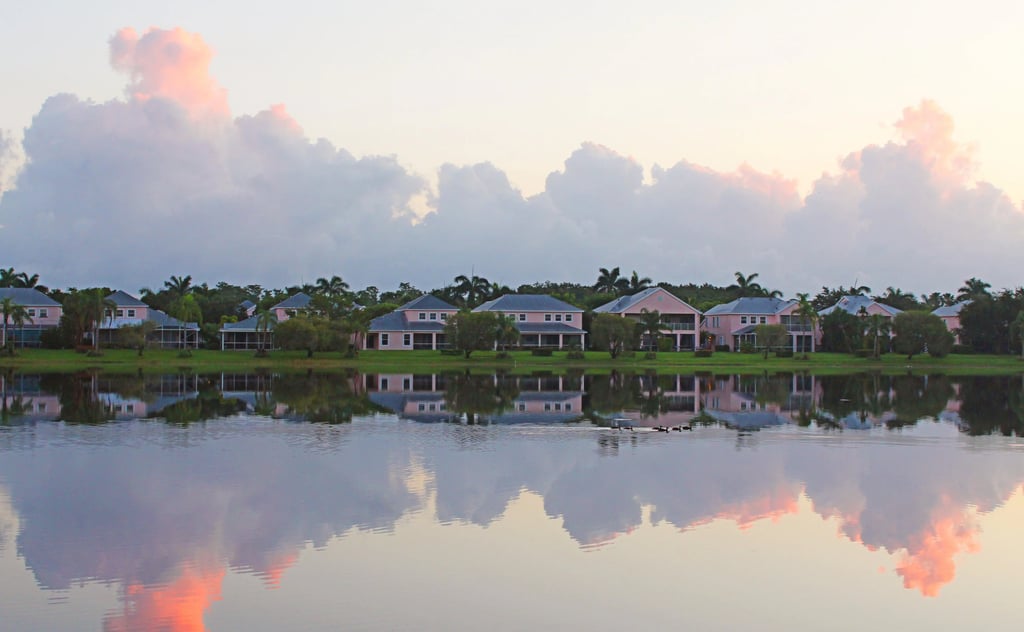 Luxury waterfront homes reflected in a calm lake under a pastel pink sunset sky with fluffy clouds.