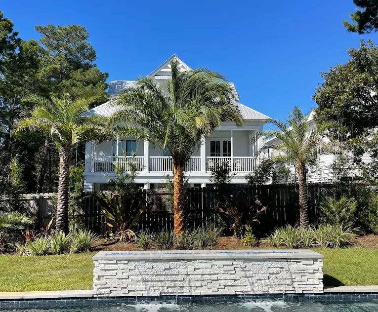 Luxury backyard swimming pool with a stone waterfall feature, palm trees, and a white beach house.