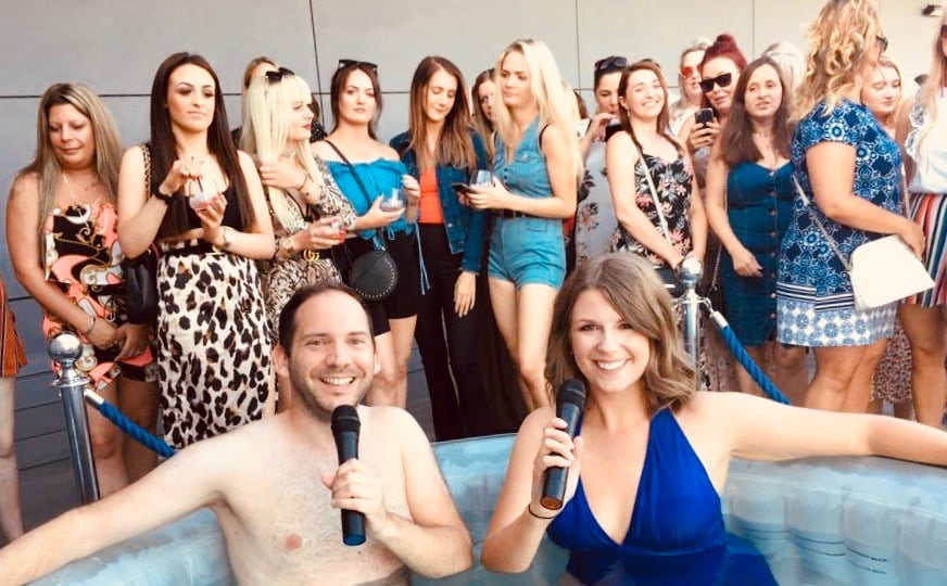 Alex Duffy and Ellie Brennan are in a hot tub with microphones, with audience behind.