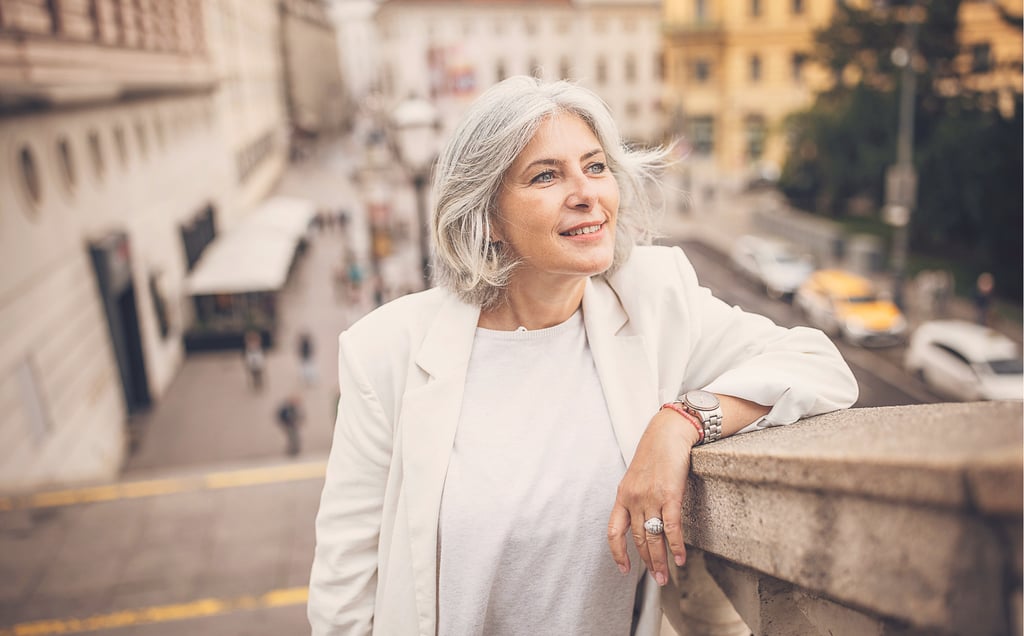 Smiling senior woman with gray hair wearing a white blazer while traveling in a European city.