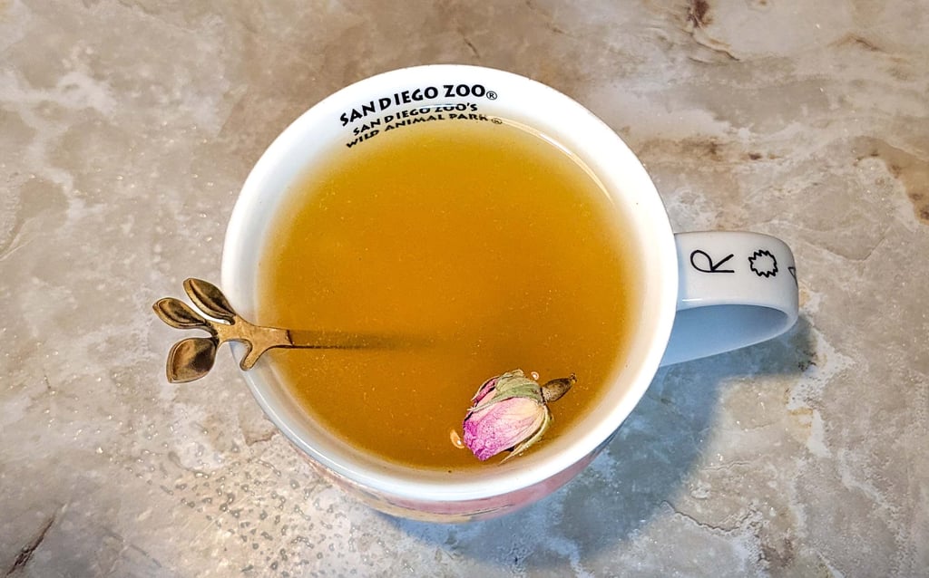 Cup of herbal tea with a small rosebud and spoon inside, placed on a table.