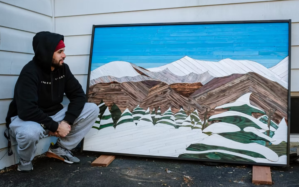 Artist displays a large reclaimed wood mountain wall art piece with snowy peaks and pine trees.