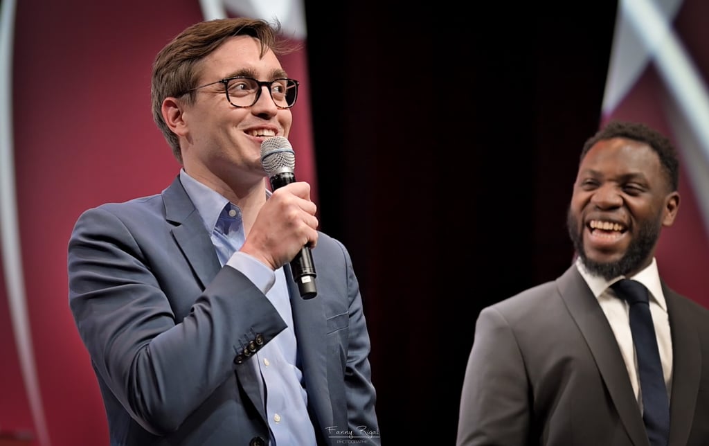 A smiling male speaker in a blue suit holding a microphone at a conference stage.