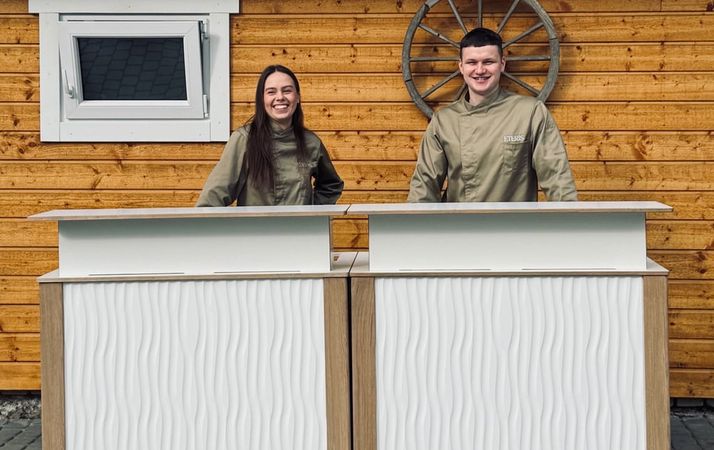 Two smiling catering staff in chef coats stand behind a modern white portable bar counter.