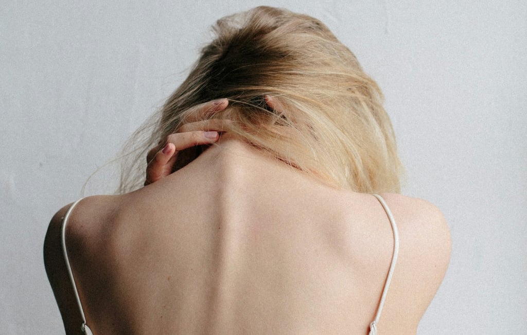 Woman with bare back and blonde hair holding her neck, symbolizing tension, pain, or vulnerability.