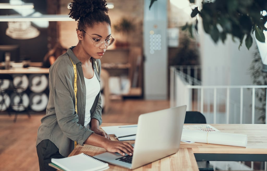 Young woman with a measuring tape typing on a notebook while standing near a desk with paperwork.