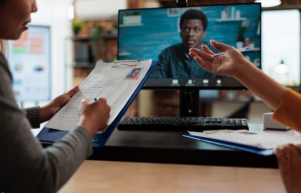 A figure looks distractedly at a CV on a clipboard whilst an interview candidate appears on screen