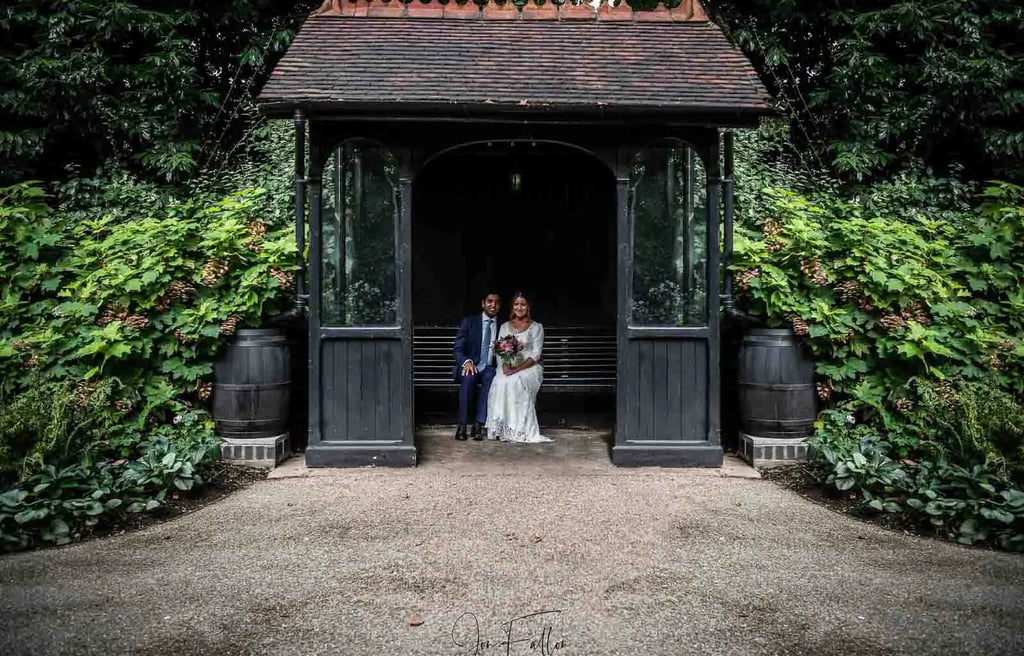 A bride and groom sitting inside a rustic garden gazebo surrounded by lush green foliage.