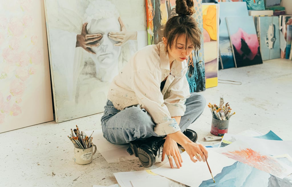 Jovem artista sentada no chão rodeada por pinturas, trabalhando em uma aquarela.