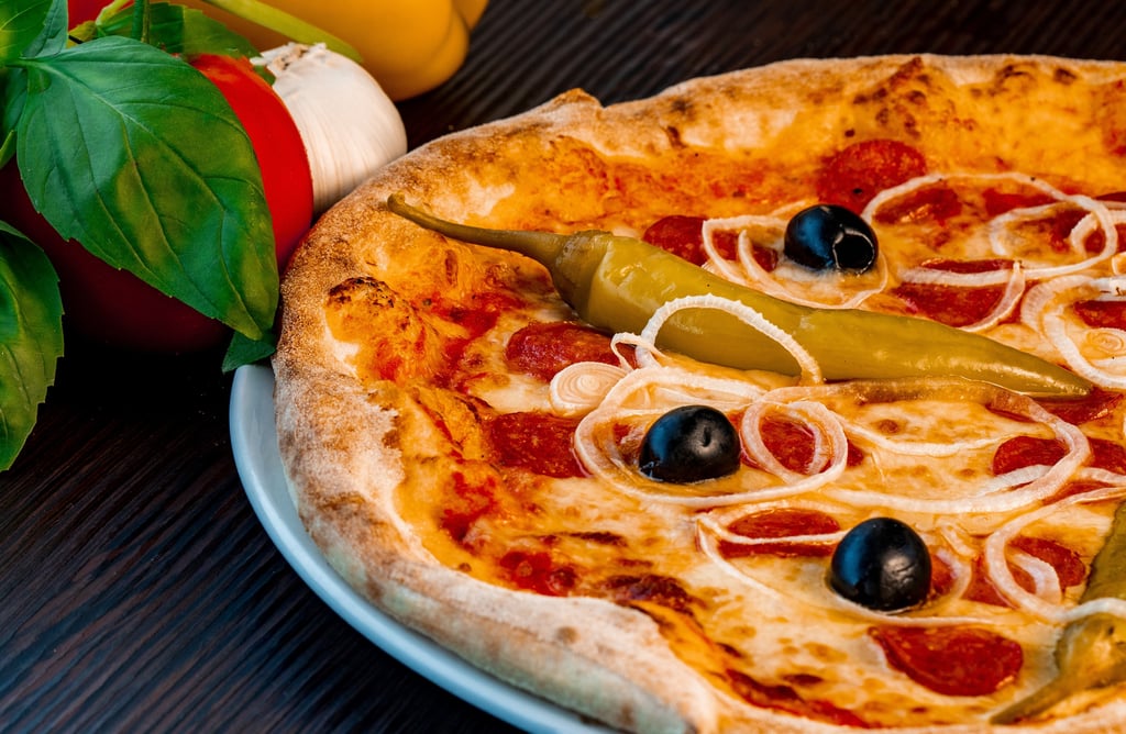 Steinofen-Pizza mit Salami, Oliven, Zwiebelringen und Peperoni neben frischem Basilikum.